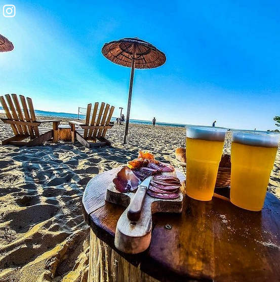 plage de sable avec chaise longue et en arrière plan la mer. Au premier plan une table avec 2 verres de boison et une planche apéro.